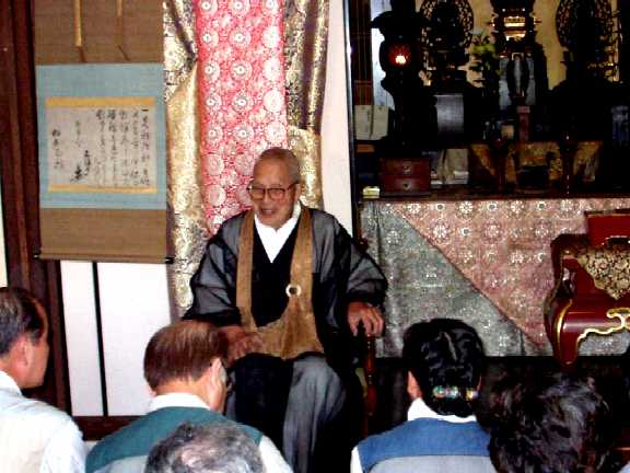 写真ａ１３：三成直筆の書と仏壇の前で講話する谷口楚碩和尚。
